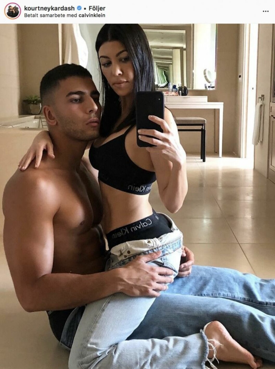 Kourtney Kardashian och Younes Bendjima har ett av-och-på-förhållande. Kourtney Kardashian sitter i Younes Bendjimas knä på golvet. Han har bara jeans på sig och hon har jeans och en sporttopp