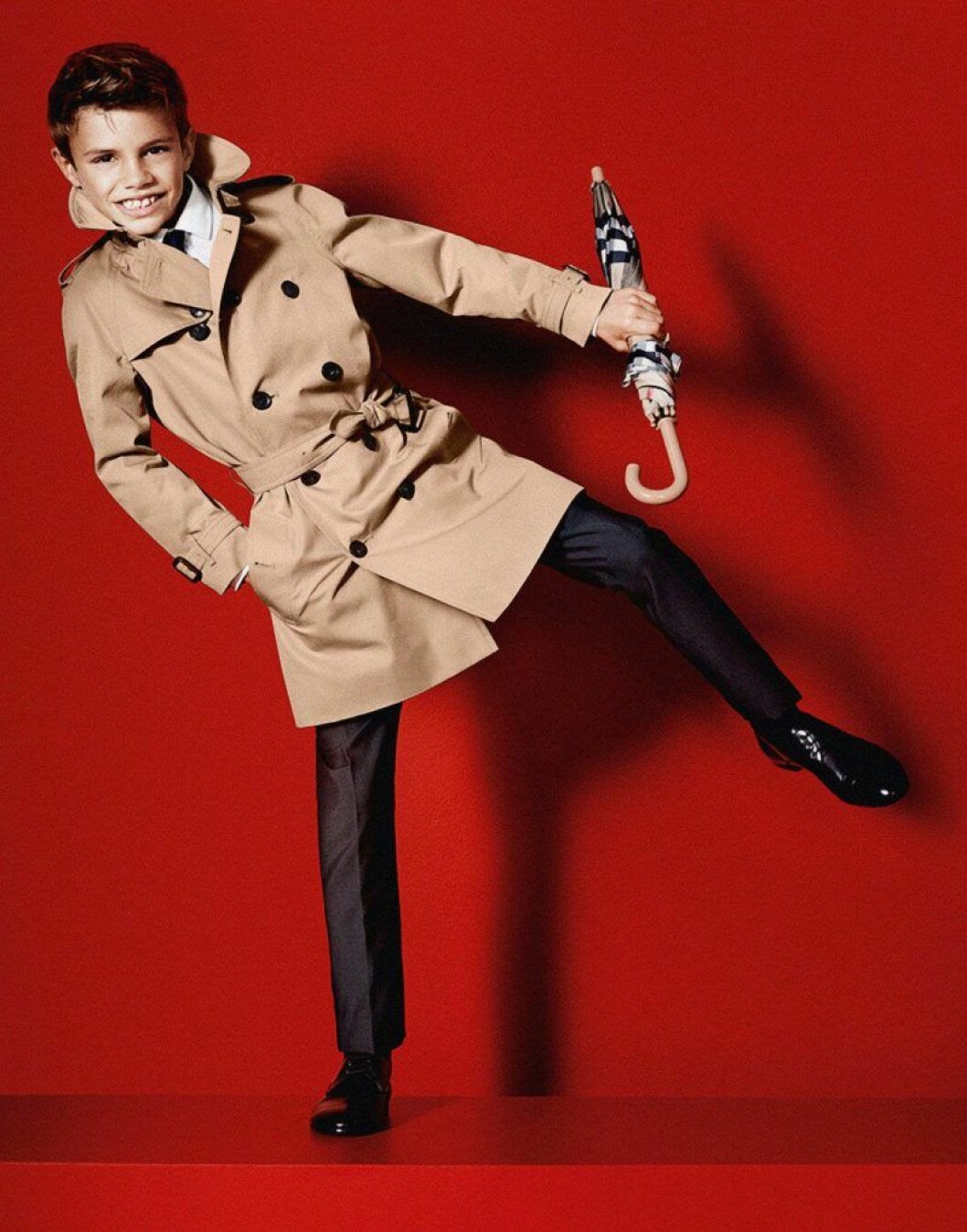 Romeo Beckham i beige kappa och paraply framför en röd bakgrund