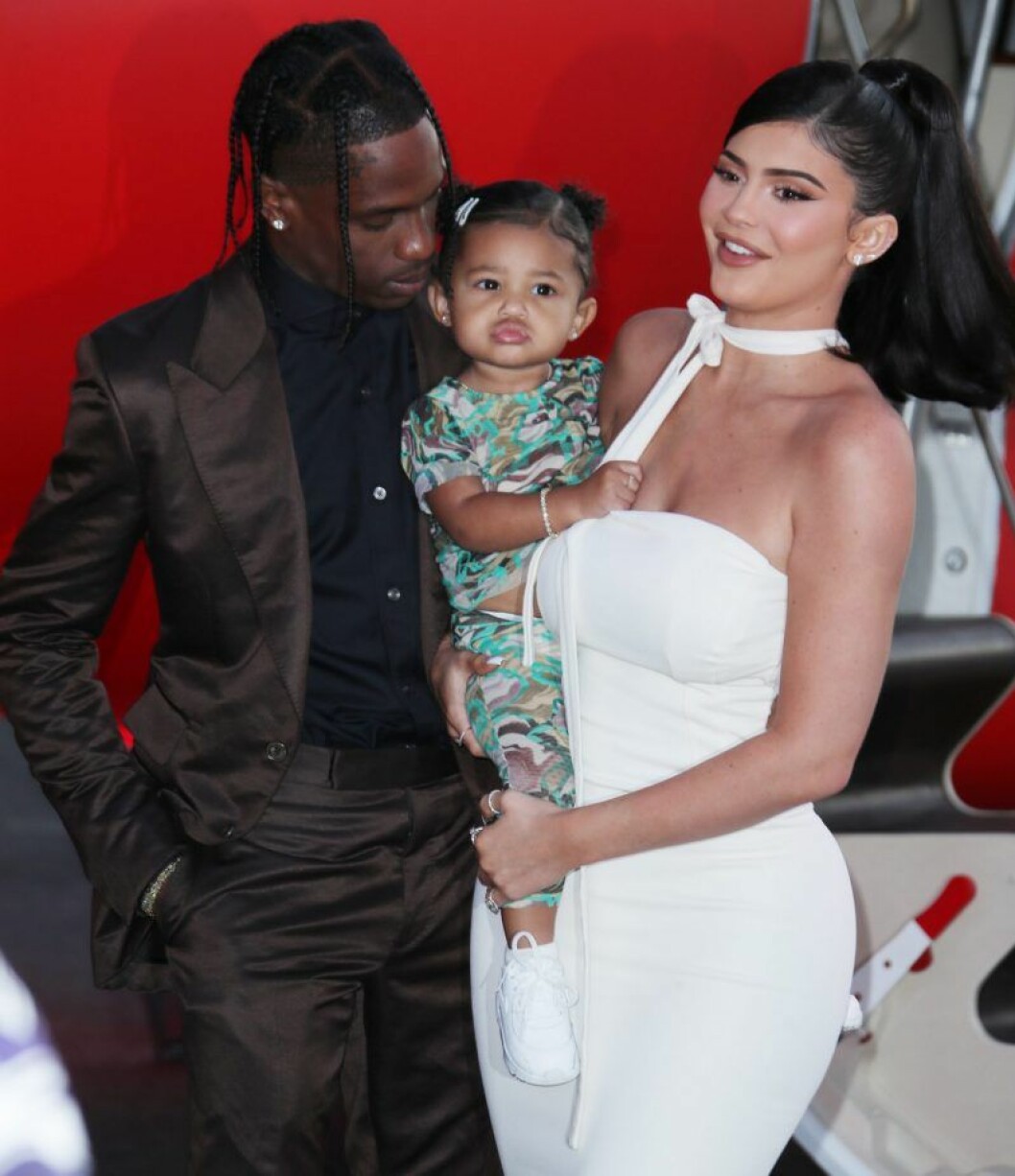 Travis Scott och Kylie Jenner tillsammans med dottern Stormi. Kylie Jenner står i vit klänning och håller dottern Stormi och Travis Scott är på väg att pussa Stormi på kinden